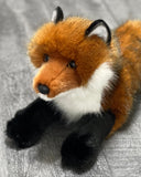 Up close view of lifelike weighted red fox plush in a laying pose with rust coat, black paws, and white ruff.
