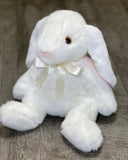 Front-side-angle view of a weighted white lap bunny plush with floppy ears, pink nose, and cream satin bow.  Ultra soft rabbit comfort companion.