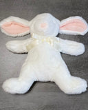 White weighted bunny plush with floppy ears and satin bow lying flat on a gray surface with arms, legs ands ears outstretched.