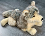 Front three-quarter view of wolf plush lying down; soft lifelike fur in grays, browns, and cream,  with a black nose visible.