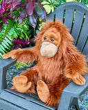 Weighted orangutan plush seated in a garden chair with arms on the armrests, tropical plants behind.