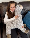 Young person sitting on a leather couch hugging an oversized weighted llama plush, showing its large cuddly body and long floppy legs.