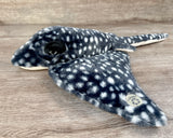 Side angle view of weighted black and white stingray plush showing a made with recycled materials tag on the wing.